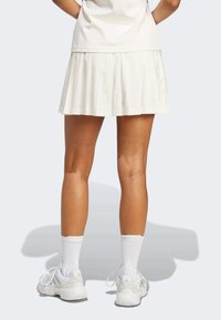 Jupe de tennis plissée blanche avec une texture lisse, assortie à des chaussures de sport blanches et des chaussettes blanches de longueur mi-mollet, vue de derrière.