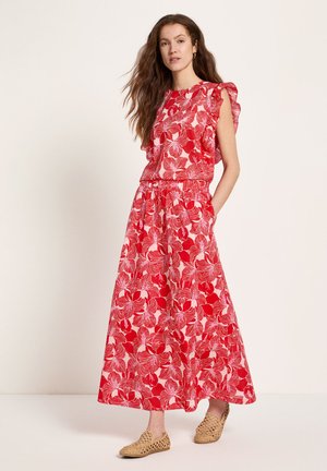 Frau mit langen Haaren, die ein rotes und rosa florales ärmelloses Oberteil und passende weite Hosen trägt, steht mit einer Hand in der Tasche.