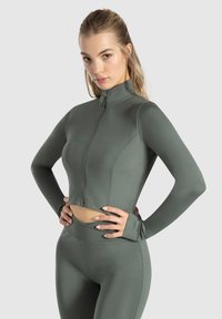 Olivenfarvet cropped atletisk jakke med lange ærmer, høj krave og frontlynlås. Glat materiale med synlige syningsdetaljer.