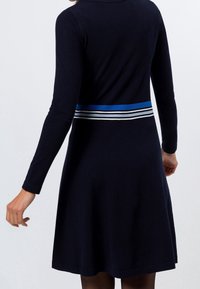 Mörkblå långärmad klänning med en figursydd bodice och utsvängd kjol, prydd med ett horisontellt randigt bälte i blått, vitt och svart vid midjan.