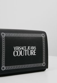 Czarny portfel z białą, sznurkowatą obwódką i napisem "VERSACE JEANS COUTURE" na środku przedniej strony.