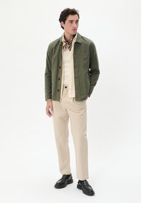 Grüne Baumwolljacke über einem cremefarbenen Hemd, kombiniert mit beigen Hosen und schwarzen Loafers. Die Jacke hat ein Knopfdesign und Taschen.