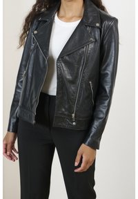 Kvinna klädd i en svart läderbikerjacka över en vit ribbad tröja och svarta byxor, står mot en enfärgad bakgrund.