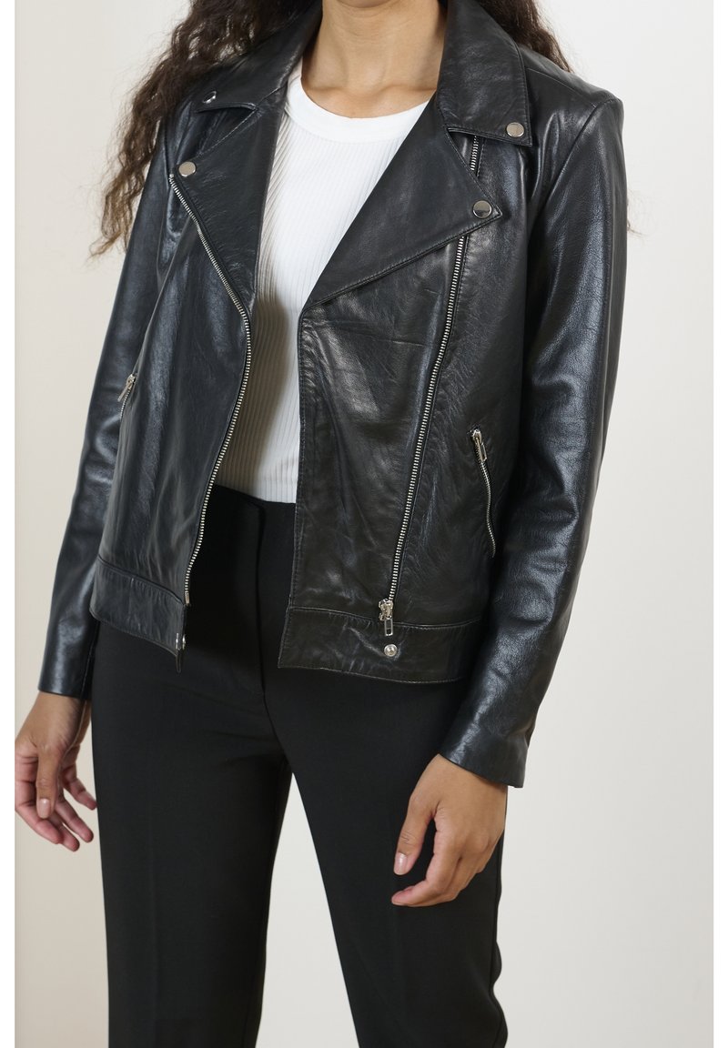 Kvinna klädd i en svart läderbikerjacka över en vit ribbad tröja och svarta byxor, står mot en enfärgad bakgrund.