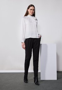 Hvit langermet bluse med høy hals og blomsterdetaljer; kombinert med svarte dressbukser og svarte ankelstøvler. Enkel, stilren design.