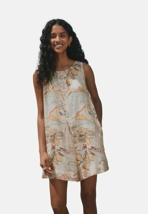 Lächelnde Frau mit langen lockigen Haaren, die ein ärmelloses Kleid über dem Knie mit Segelboot-, Zitronen-, Blumen- und Anker-Motiven auf hellem Hintergrund trägt.