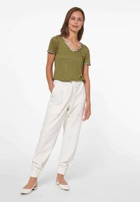 Olijfgroene t-shirt met een V-hals en een patroonrand, gecombineerd met witte joggingsbroeken met elastische boorden en zakken. Witte flats.