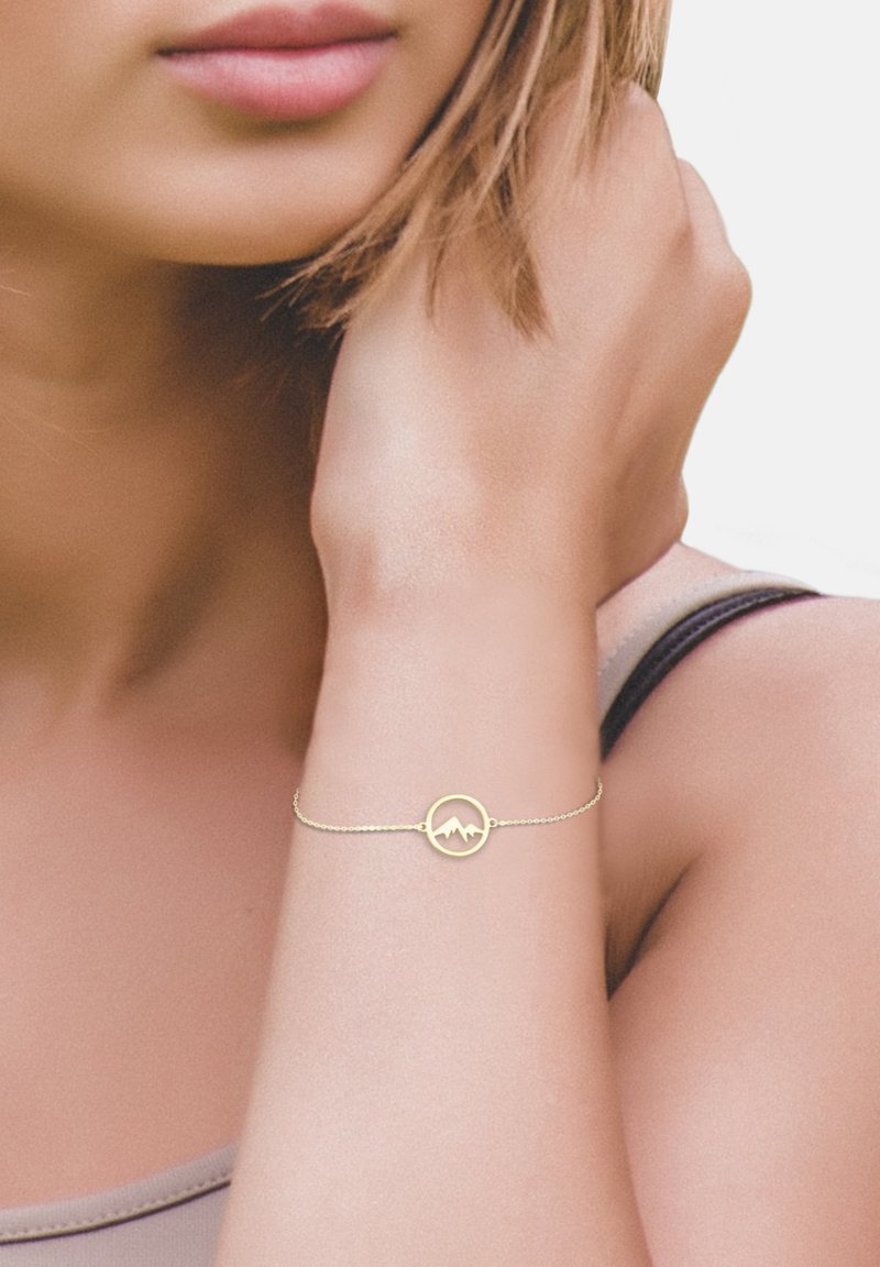 Bracelet en or avec un pendentif circulaire arborant un motif de montagne ; chaîne délicate, style minimaliste, porté au poignet. Teint de peau et vêtements partiellement visibles.