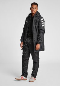Schwarze gepolsterte Jacke mit Kapuze, elastischen Bündchen und weißen Schulterstreifen. Kombiniert mit schwarzen Hosen und grauen Schuhen mit roten Akzenten.