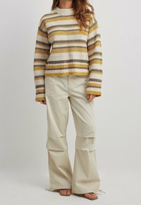 Raidoitettu neulepaita keltaisena, beige ja ruskea; lyhyt malli, pitkät hihat. Beiget cargo-housut, joissa on leveät lahkeet ja narukiinnitykset.