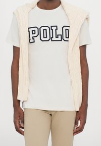 Bílý bavlněný tričko s nápisem "POLO" černými písmeny, vrstvené pod krémovým svetr-vestou s copánkovým vzorem. Béžové elegantní kalhoty.