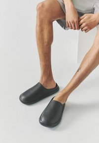 Svarta slip-on-skor i slätt material, med en rundad tådesign och en minimalistisk estetik. Bärs med neutrala shorts.