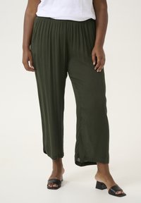 Tummanvihreät, laskostetut leveät housut joustovyöllä, yhdistettynä mustiin neliönmuotoisiin korokepohjaisiin sandaaleihin neutraalilla taustalla.