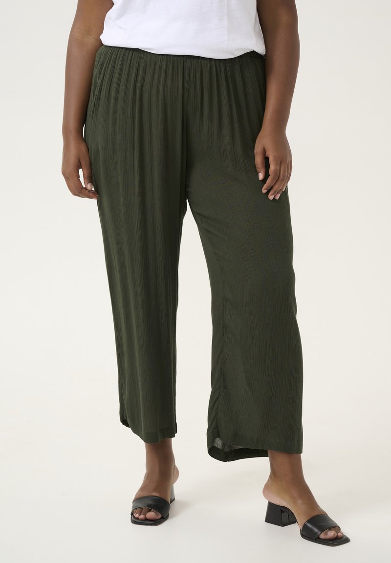 Tummanvihreät, laskostetut leveät housut joustovyöllä, yhdistettynä mustiin neliönmuotoisiin korokepohjaisiin sandaaleihin neutraalilla taustalla.