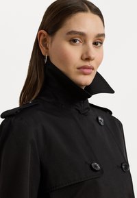 Casaco preto de trench com um colarinho alto, apresentando botões duplos e epaulettes nos ombros. Textura suave e design ajustado.