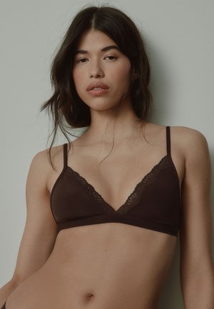 Jeune femme aux longs cheveux foncés portant un bralette marron foncé orné de dentelle sur un fond clair uni.