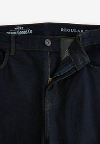 Donkerblauwe spijkerbroek met een open ritssluiting, knoop dicht, oranje stiksels en tekst op de tailleband met de tekst "Next Denim Goods Co Regular".