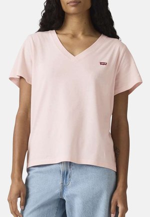 Femme portant un t-shirt rose clair à col en V avec un petit logo Levi's rouge et un jean bleu clair, debout devant un fond uni.