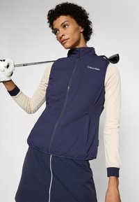 Colete azul-marinho com fecho em zip, bolsos laterais e barra elástica. Combinado com uma blusa de manga longa em creme com punhos canelados.