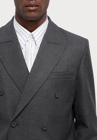 Giacca da abito grigia doppiopetto con finitura testurizzata, dotata di revers a punta e una tasca sul petto, indossata sopra una camicia a righe bianco e blu.