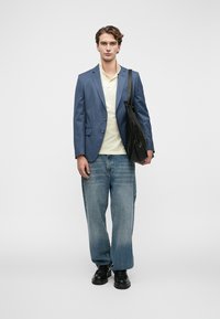 Νεαρός άνδρας που φοράει μπλε σακάκι, κρεμ πόλο, φαρδιά μπλε τζιν, μαύρα παπούτσια, κρατώντας μαύρη τσάντα ώμου, στέκεται στραμμένος προς τα εμπρός.