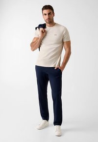T-shirt beige ajusté avec logo, pantalon marine et baskets blanches. Le modèle tient un pull marine sur son épaule, debout devant un fond clair.