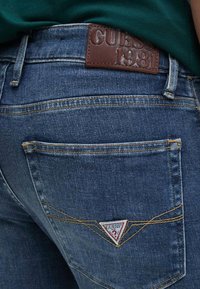 Jeans de mezclilla en azul oscuro con una textura suave, que presentan un parche de cuero marrón grabado con "GUESS 1981" y un logo en forma de triángulo en el bolsillo trasero.