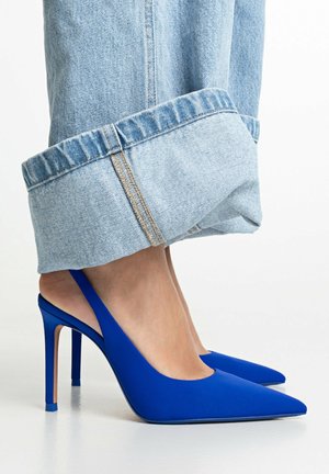 Pies con tacones altos slingback de punta en azul brillante combinados con vaqueros azules claros con puños, sobre un fondo sencillo.