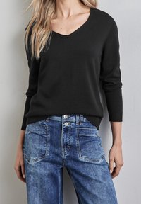 Svart stickad tröja med V-hals, bärs över ljusblå denimjeans med en urtvättad finish och silverfärgade knapppdetaljer. Texturerad tyg synlig.