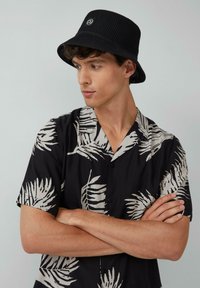 Black and white patterned short-sleeve shirt with palm leaves, buttoned down, paired with a black textured bucket hat featuring a logo.