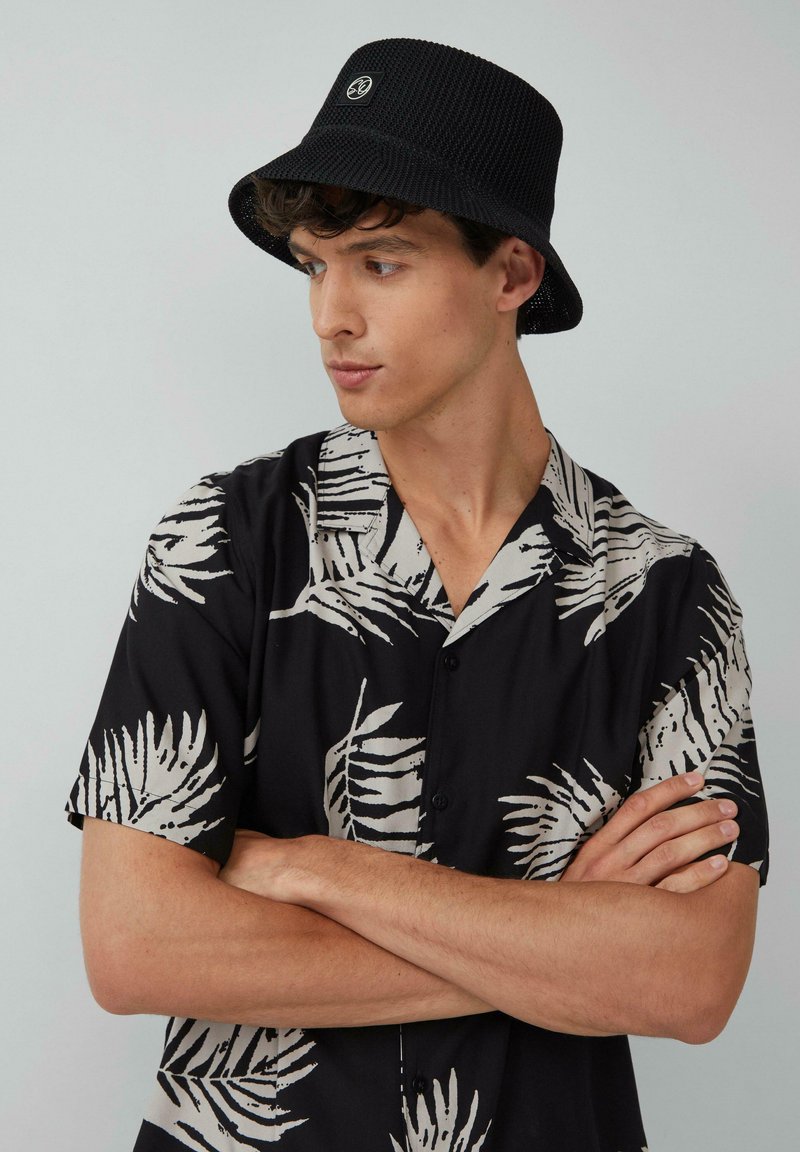 Black and white patterned short-sleeve shirt with palm leaves, buttoned down, paired with a black textured bucket hat featuring a logo.