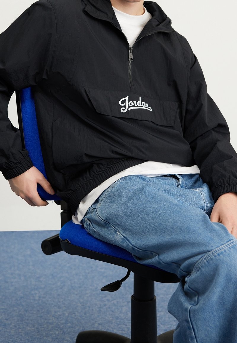 En person sitter på en blå kontorsstol, iklädd en svart Jordan zip-up jacka och ljusblå jeans, med händerna vilande på låren och armstödet.