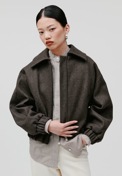 Giubbotto bomber in misto lana marrone con colletto alto, chiusura con zip e polsini arricciati, indossato sopra una camicia a righe con bottoni.