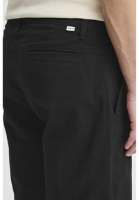 Pantaloni dintr-un amestec de bumbac negri, cu un design drept, buzunare laterale și un buzunar posterior cu o etichetă vizibilă. Textură netedă.