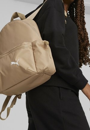 Osoba nosząca beżowy plecak Puma z białym logo, ubrana w czarne długie rękawy i spodnie, z widocznymi długimi kręconymi włosami.