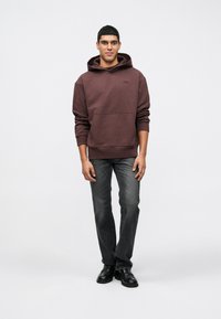 Άντρας που στέκεται με τα χέρια χαλαρά, φορώντας ένα μπορντό φούτερ, ξεθωριασμένο μαύρο τζιν και μαύρα παπούτσια, μπροστά σε απλό φωτεινό φόντο.