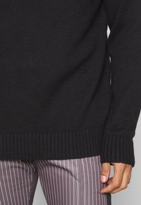 Schwarzer gestrickter Pullover mit gerippten Bündchen und Saum. Verfügt über ein strukturiertes Design und eine lockere Passform, kombiniert mit gestreiften helllilanen Hosen.