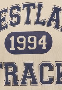 Tela crema con texto en azul marino que dice "WESTLAND TRACK 1994". El texto es en negrita, con una textura ligeramente elevada alrededor de las letras.