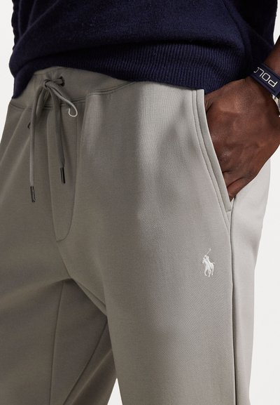 Svetlo šedé tepláky s elastickým pásom a šnúrkou, s bočnými vreckami a bielou vyšitou značkou na ľavej nohe.