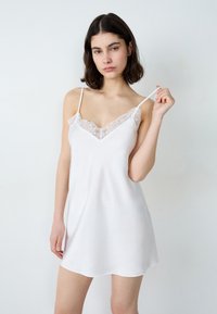 Robe nuisette en satin blanc avec des bretelles spaghetti réglables et un col en dentelle, offrant une texture douce et lisse ainsi qu'une silhouette fluide.