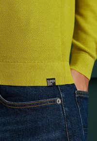 Pull en maille jaune vif avec motif texturé, poignets et ourlet côtelés. Étiquette en bas à gauche indiquant "SUPERDRY." Associé à un jean denim foncé.