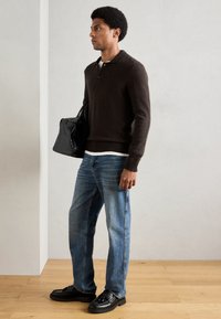 Σκούρος καφέ πλεκτός σκούφος με γιακά, μπλε ίσια τζιν παντελόνια και μαύρα δερμάτινα παπούτσια. Κρατώντας μια μαύρη τσάντα με υφή.