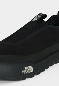 Baskets noires sans lacets avec dessus texturé et semelle épaisse, comportant le logo The North Face sur le côté et une languette.