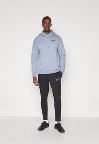 Svetlo modrá mikina s kapucňou a predným vreckom, čierny nápis "SPURS" a logo Nike. Spárované s čiernymi úzkymi teplákmi a čiernymi športovými topánkami.