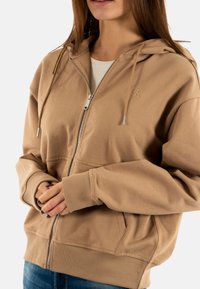 Veste à capuche beige à zip, avec poches kangourou et poignets côtelés. Présente un logo discret sur la poitrine et une fermeture éclair en métal.