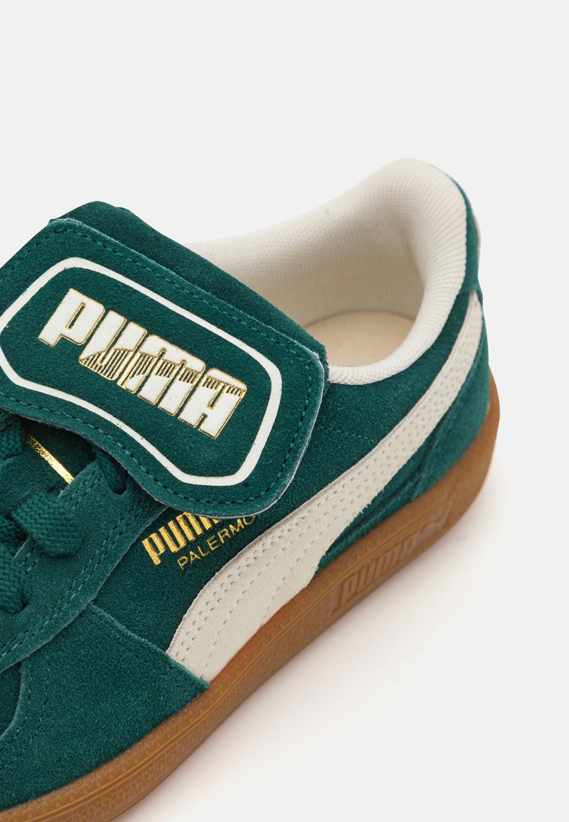 Sneaker in suede verde con pannello bianco, dettagli dorati e una grande fascetta in velcro con il nome del marchio. Suola in gomma visibile nella parte inferiore.