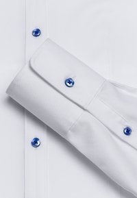 Chemise blanche en tissu lisse, avec des boutons bleus et des poignets repliés pour une touche de détail. Design de col classique, look propre et ajusté.