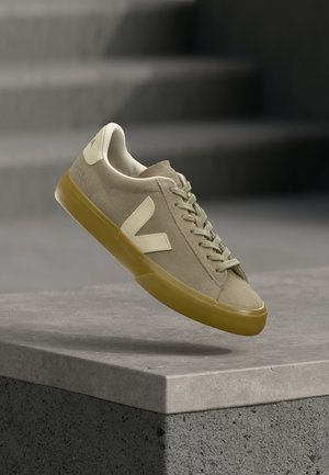 Lichtbeige suède sneaker met crème logo en veters, mosterdgele zool, zwevend weergegeven boven een betonnen oppervlak met vervaagde grijze trappen op de achtergrond.