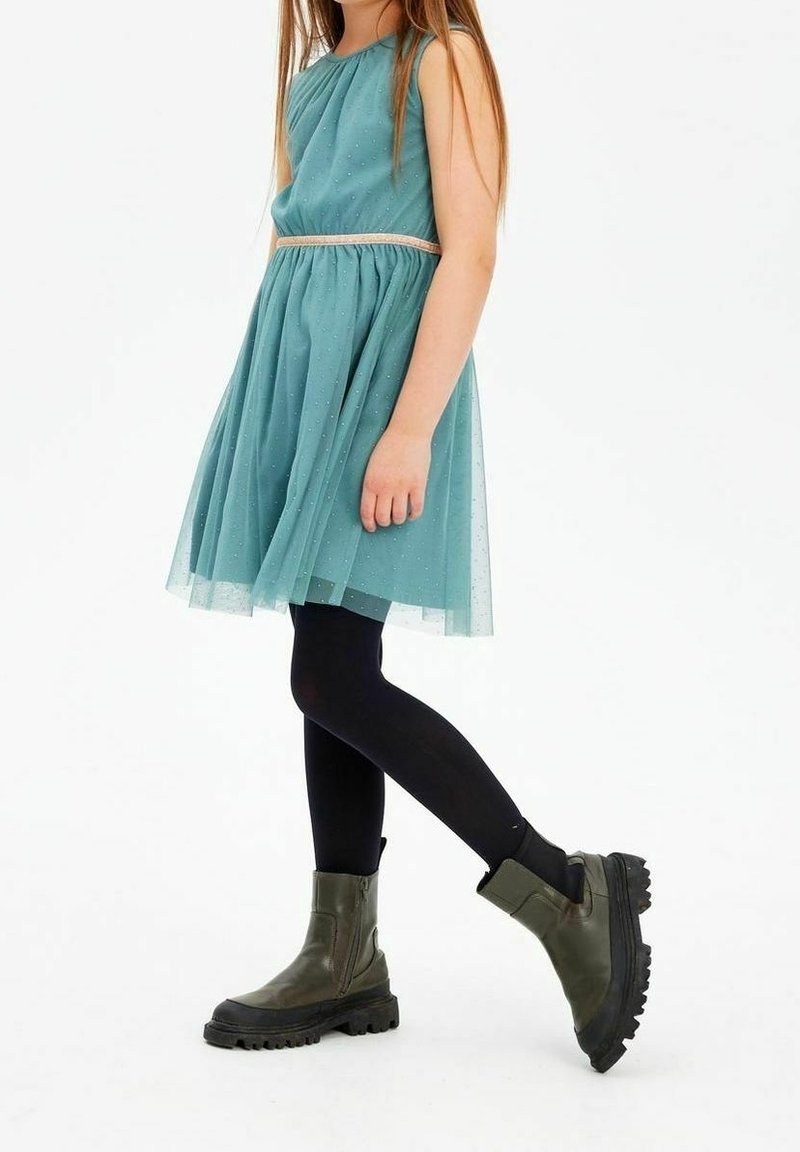Enfant portant une robe sans manches turquoise avec une ceinture beige, des collants noirs et des bottes épaisses vert olive à semelles épaisses sur fond blanc.