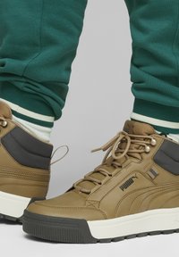 Bruna läderhögtoppsneakers med svarta accenter, gummisula och beige snören, kombinerat med gröna sweatpants med vita ränder.